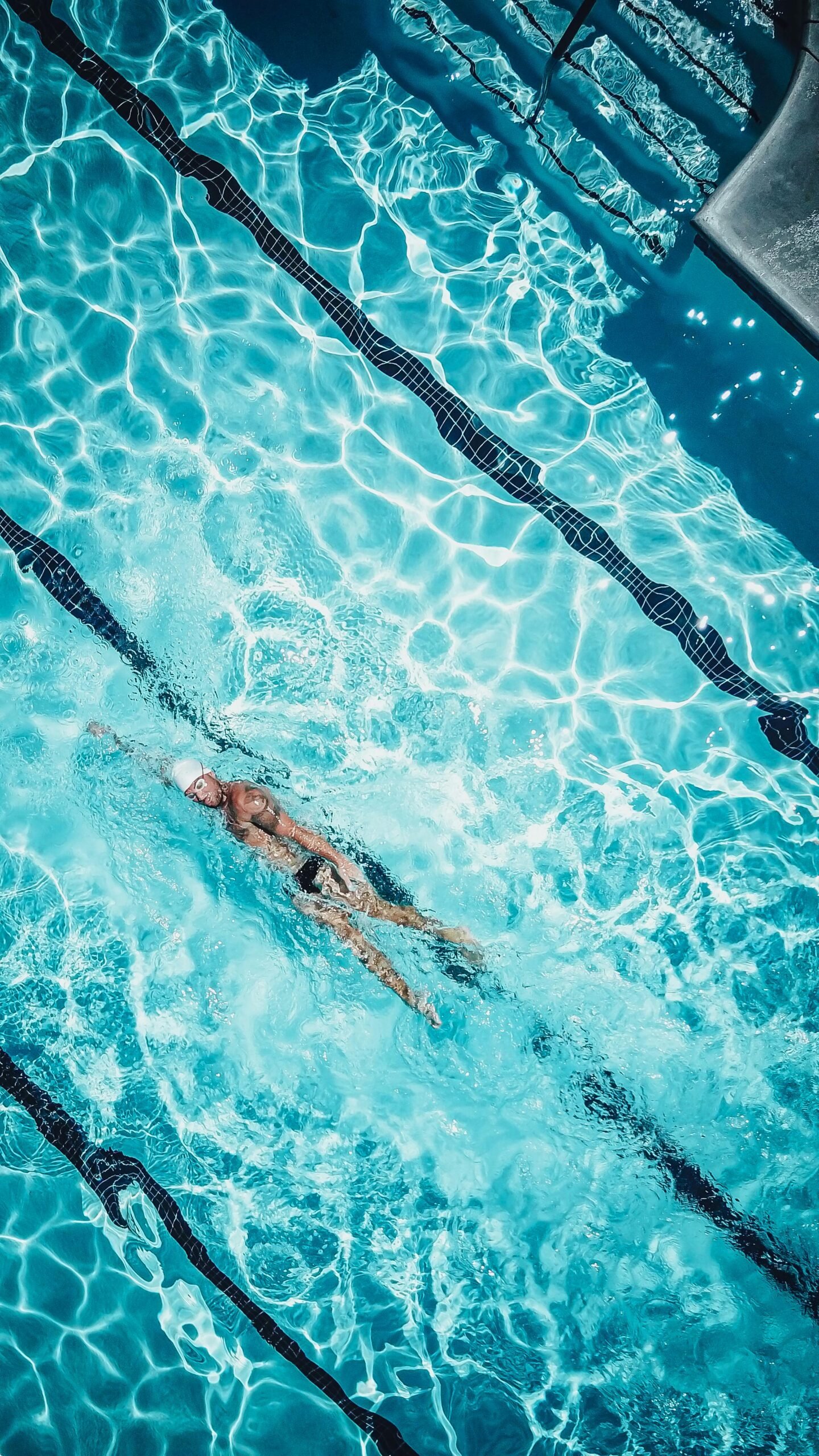 A man swimming in a pool, his arms stretched forward in a front crawl stroke, creating ripples in the clear blue water. Sunlight reflects off the surface, highlighting his steady movement and the calm surroundings of the pool.